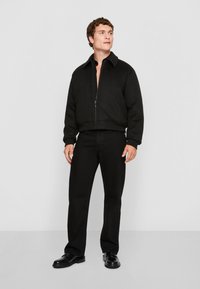 Chaqueta negra corta con cuello alto y cremallera frontal, combinada con jeans negros y zapatos negros pulidos, sobre un fondo blanco.