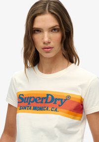 Jonge vrouw met golvend bruin haar draagt een wit SuperDry T-shirt met oranje en rode strepen en blauwe tekst met de woorden "SuperDry Santa Monica, CA."