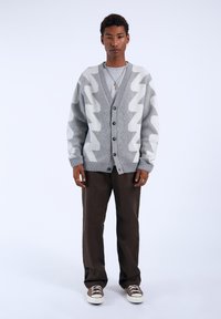Gray and white zigzag patterned cardigan, buttoned up, with ribbed cuffs. Paired with brown trousers and light sneakers, standing on a plain background.