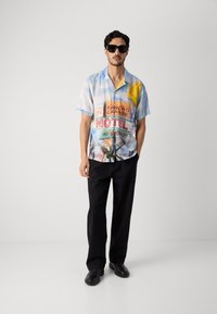 Majica s kratkimi rokavi svetlo modre barve s pisanem natisom motela, ki vključuje palme in sonce, v kombinaciji s črnimi hlačami in čevlji.