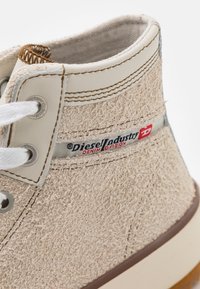 Beige textile ankle boot featuring a fuzzy finish, leather accents, white laces, and a logo patch on the side, with a brown rubber sole.