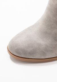 Bota de ante gris hasta el tobillo con punta puntiaguda, que presenta una textura suave y una suela en tono cuero, destacando elementos de diseño minimalista.