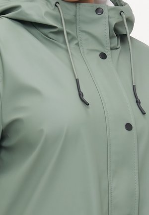Gros plan d'un imperméable vert clair avec des boutons-pression noirs et des cordons sur la capuche.