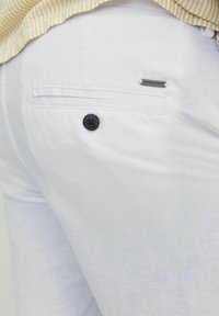 Pantalones de lino blancos con un bolsillo trasero, que cuenta con un botón negro y una pequeña etiqueta de metal con logotipo. La tela tiene una textura ligera.