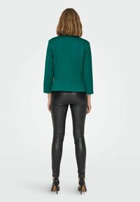 Vihreä räätälöity jakku, jossa on sileä pinta ja kolme neljänneksen hihaa, yhdistetty mustiin istuviin nahkahousuihin ja korkeakorkoisiin kenkiin.