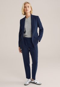 Traje de color azul marino a medida con un corte slim, que incluye un blazer de un solo botón y pantalones frontales planos, combinado con un suéter de punto gris y zapatillas blancas.