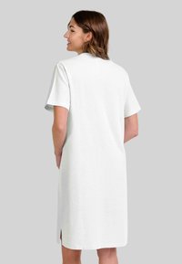 Mujer con cabello castaño que lleva un vestido blanco de manga corta hasta la rodilla, de pie y sonriendo de perfil sobre un fondo neutro.