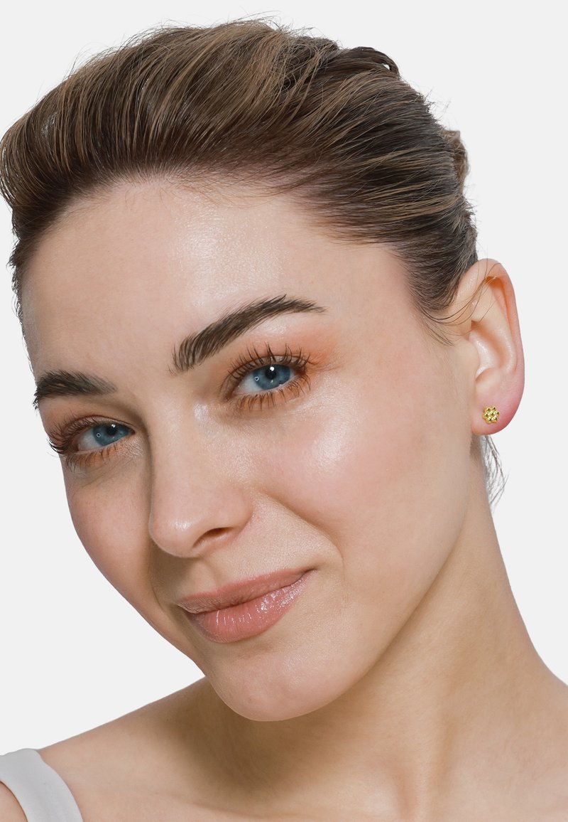 Boucles d'oreilles en or avec un design floral. Surface lisse avec de petites pierres précieuses. Cheveux tirés en arrière, mettant en valeur les boucles d'oreilles.