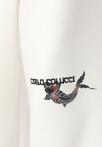 Hvit stoff med et Koi-fisk grafikk i svart, rødt og hvitt, med "CARLO COLUCCI" tekst brodert under designet.