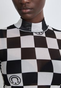 Sheer black and white checker-patterned top with a high collar and black trim. Features a circular logo in the lower right.