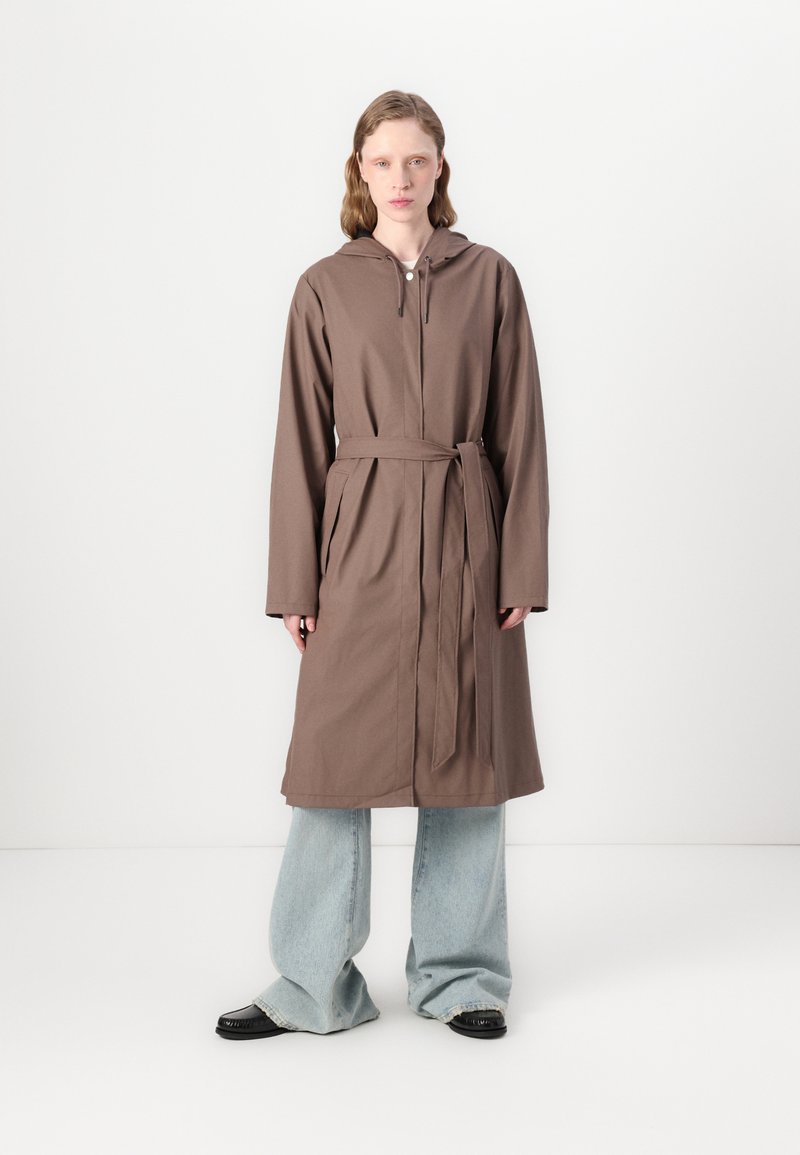 Brown, hooded, long-sleeve coat with a tie belt. The fabric appears smooth; model wears blue wide-leg jeans and black shoes.