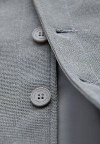 Gros plan d'un tissu gris texturé avec deux boutons ronds gris, un cousu et un partiellement visible, sur une ouverture de vêtement.