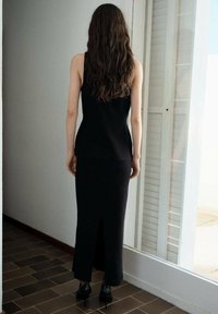 Haut noir sans manches associé à une longue jupe noire avec une fente arrière, porté avec des chaussures noires à talons hauts. Tissu lisse, design ajusté.