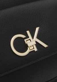 Superficie in pelle nera testurizzata con un emblema dorato metallico "CK", caratterizzata da una piccola chiusura rettangolare che collega le lettere.