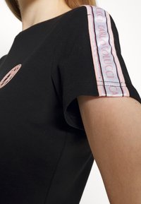 Czarna koszulka z okrągłym dekoltem. Posiada różowy haftowany logo z przodu oraz różowo-niebieską taśmę w paski na ramieniu.