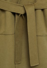 Oliivinvihreä, pehmeäpintainen kangas, jossa näkyy solmittava vyö; varustettu kahdella suorakulmaisella taskulla. Kangas näyttää huovutetulta ja lämpimältä.