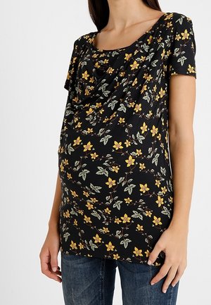 Zwangere vrouw draagt een zwarte zwangerschapsblouse met gele bloemenprint en korte mouwen, gecombineerd met een blauwe spijkerbroek.