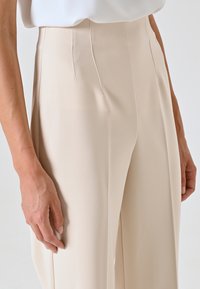 Beige wijde broek met een gladde textuur, voorzien van een hoge taille, subtiele plooi en een vlakke voorkant. Hand rustend op de stof.