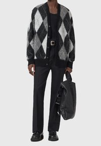 Personne portant un pantalon noir, une chemise noire, un cardigan noir et blanc à motif losange, des chaussures noires, et tenant un sac à dos en cuir noir dans une main.