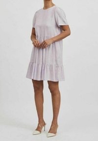 Žena v krátkých rukávech, světle fialových šatech ke kolenům s volánkovým lemem a bílých špičatých lodičkách na vysokém podpatku.