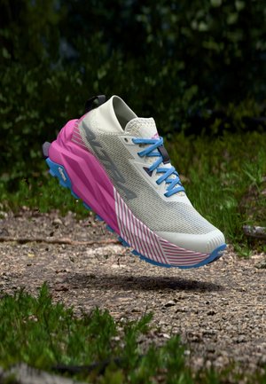Leichtgrauer, rosa und blauer Trailrunning-Schuh in der Luft über einem Kiesweg mit grünem Laub im Hintergrund.