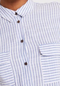 Koszula w niebiesko-białe paski z kołnierzykiem, z zapięciem na guziki i dwoma przednimi kieszeniami o różnych wzorach pasków. Miękka tekstura.