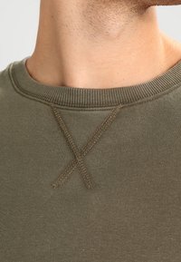 Suéter verde oliva de cuello redondo con una tela texturizada, cuello acanalado y un detalle de costura en forma de X cerca del cuello.