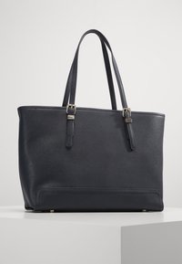 Sac en cuir noir avec de longues sangles réglables et des détails de boucle en ton or, exposé sur un piédestal blanc.