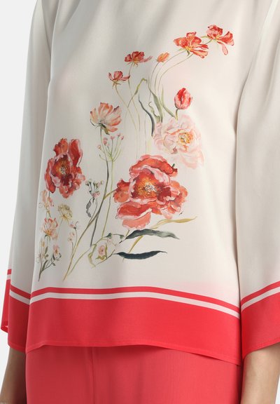 Blouse blanche à imprimé floral rouge et rose avec un détail d'ourlet rouge au-dessus d'un pantalon rouge corail, montrant la taille et le bras.