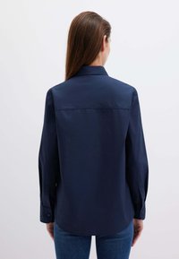 Camicia blu navy a maniche lunghe con bottoni, orlo dritto e yoke posteriore. Il tessuto appare liscio, con polsini abbottonati e un design del colletto classico.