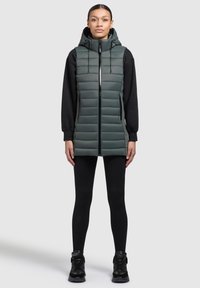 Vadderad väst i mörkgrön med huva, dragkedja fram och sidofickor. Bärs över en svart långärmad topp och svarta leggings. Strukturerad yta.