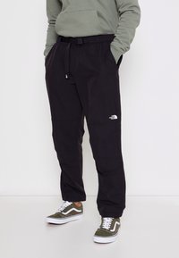 Osoba v černých kalhotách North Face s nastavitelným pásem a olivově zelených teniskách Vans, s rukama v kapsách, stojí na bílém pozadí.