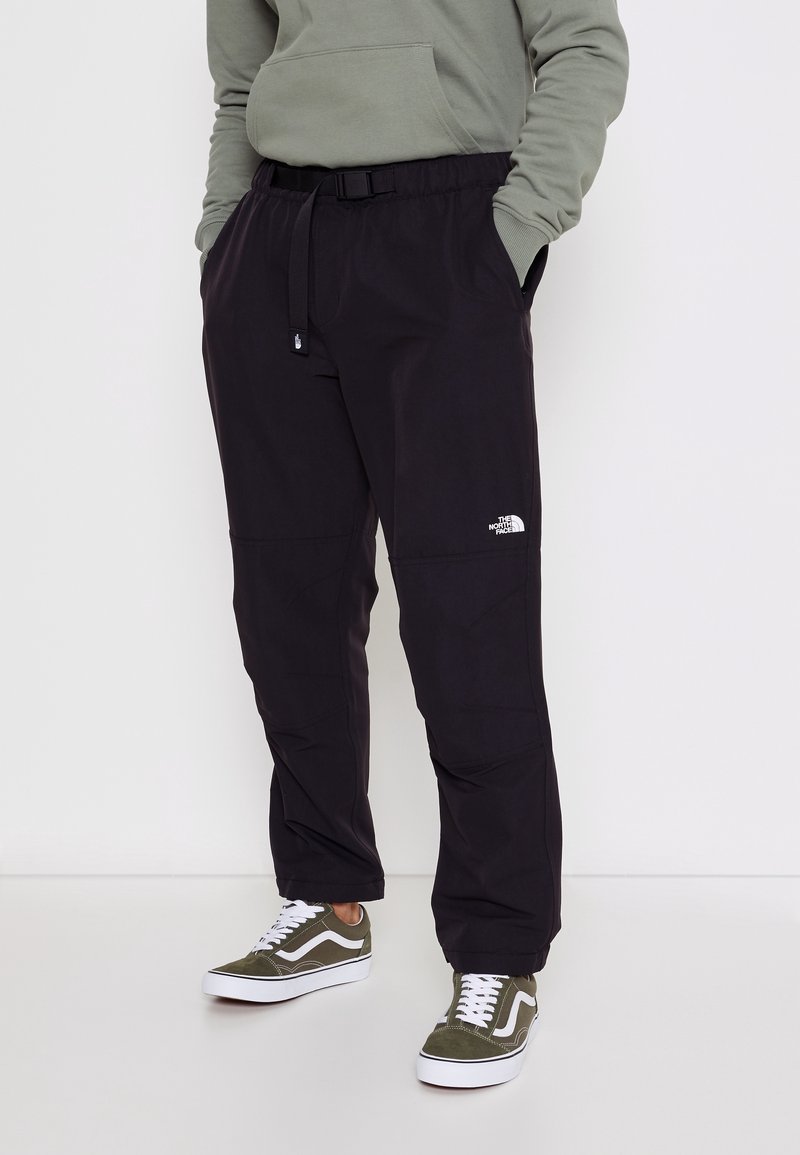 Osoba v černých kalhotách North Face s nastavitelným pásem a olivově zelených teniskách Vans, s rukama v kapsách, stojí na bílém pozadí.