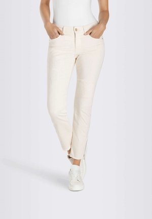 Kobieta ubrana w dopasowane, ecru jeansy ze strzępionymi nogawkami i białe trampki, z rękami w kieszeniach, stojąca na jednolitym tle.