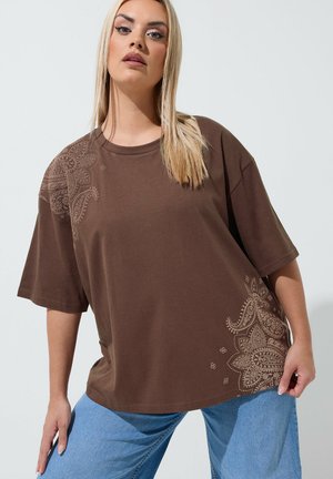 Młoda kobieta o prostych blond włosach, ubrana w luźny brązowy T-shirt z wzorami paisley oraz niebieskie dżinsy na jednolitym tle.