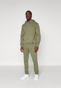 Olivza zaļš džemperis ar kangaroo kabatu un auklām, kombinēts ar atbilstošām treniņbiksēm. Valkāts ar baltām sporta apaviem ar gludu virsmu.