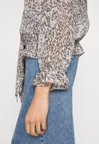 Gennemsigtig, let bluse med brun og grå dyreprint. Har rynkede ærmer og en løs pasform, parret med blå denimjeans.