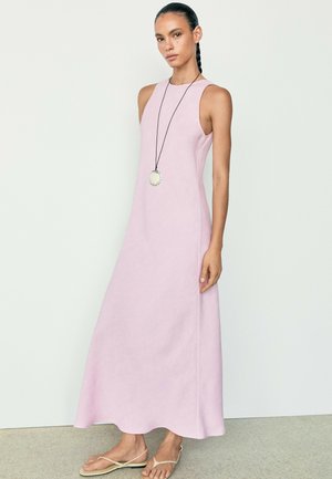 Donna con i capelli intrecciati che indossa un vestito lungo rosa chiaro senza maniche, una lunga collana e sandali bassi beige su uno sfondo chiaro e uniforme.