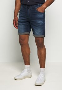 Farkkuhousut, joissa on joustava vyötärö ja naru, tummansininen väri, taitettu helma ja sivutaskut, yhdistettynä valkoisiin lenkkareihin.