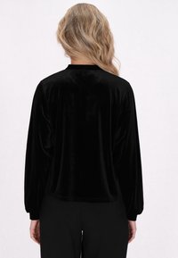 Top lungo in velluto nero a maniche lunghe con vestibilità comoda, scollo rotondo e spalle cadenti, mostrato da dietro. Texture liscia e design leggero.