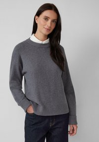Vrouw met lang, donker haar die een grijze gebreide trui over een witte blouse en donkere jeans draagt, staande tegen een effen lichte achtergrond.