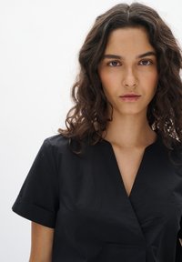 Camicetta nera a maniche corte con scollo a V e colletto ripiegato. Realizzata in tessuto leggero, presenta una leggera lucentezza e un design minimalista.