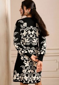 Čierne šaty s bielym kvetinovým vzorom, dlhé rukávy a rebrované manžety. Jednofarebný čierny chrbát s elegantnou, detailne spracovanou pletenou textúrou.