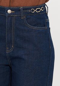 Jean en denim bleu foncé avec une accent chaîne argentée sur la ceinture, des coutures dorées et un design classique à cinq poches.