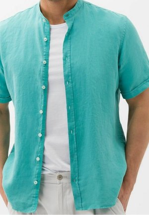 Homme portant une chemise turquoise à manches courtes ouverte sur un t-shirt blanc avec un pantalon beige, les mains dans les poches.