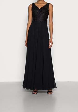 Hihaton musta iltapuku pitsisellä yläosalla ja V-pääntiellä, laskeutuva sifonkihame, käytetty mustien avokkaiden kanssa harmaalla lattialla.