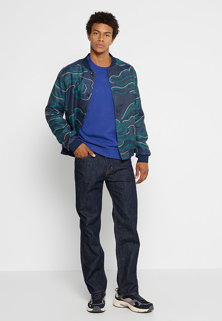 Veste de bombardier marine avec des motifs abstraits verts, associée à un sweatshirt bleu à col rond, un jean sombre et des baskets noir et blanc.