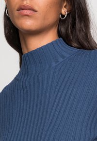 Hedvábný pletený rolák v tmavě námořnické barvě, který má vysoký límec a hladkou texturu, s viditelnými stříbrnými kruhovými náušnicemi.