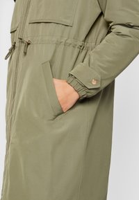 Olivově zelená parka se zipem, elastickými manžetami, nastavitelným stahováním v pase a dvěma bočními kapsami; hladká, matná textura.