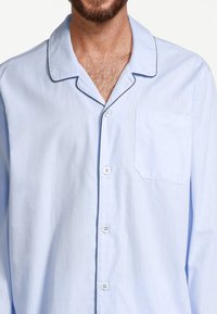 Vaaleansininen puuvillapaitapusero, jossa on kaulus, nappikiinnitys, yksi rintatasku ja tummat kontrastiväriset kaitaleet kauluksen reunoilla.